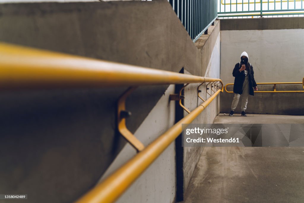 Woman wearing hooded jacket and protective face mask using mobile phone in underground