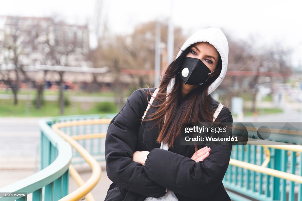 Woman wearing face mask and hooded jacket staring while standing with arms crossed