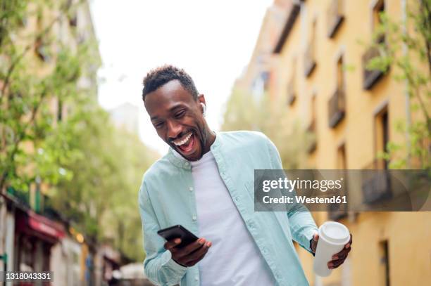 laughing young man with reusable cup using smart phone at street - bluetooth stock-fotos und bilder