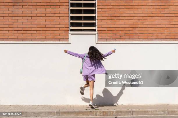 mid adult woman with arms outstretched dancing in front of wall during sunny day - woman front and back stockfoto's en -beelden