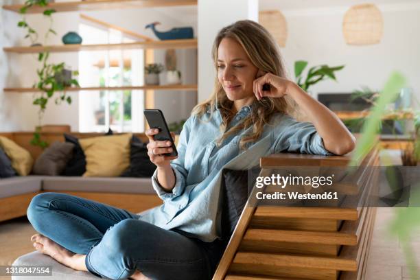 blond hair woman text messaging through smart phone while sitting in living room - smart phone woman stock-fotos und bilder