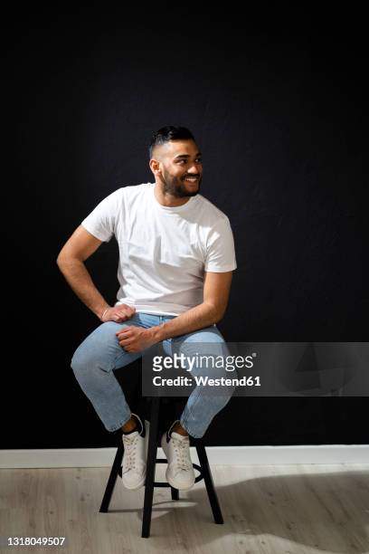 smiling man looking away while sitting on stool by black wall - style studio day 1 stock-fotos und bilder