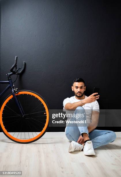 young man with smart phone sitting near bicycle by wall at studio - sitting and using smartphone studio stock-fotos und bilder