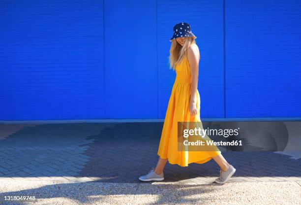 woman walking on footpath by blue wall during sunny day - long bright yellow dress stock pictures, royalty-free photos & images