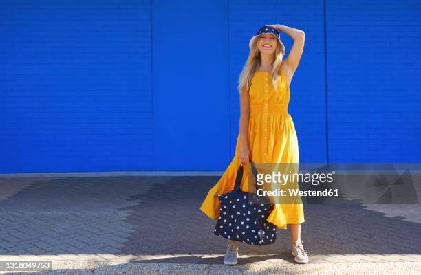 smiling woman with duffel bag standing in front of blue wall on sunny day - bucket hat stock pictures, royalty-free photos & images