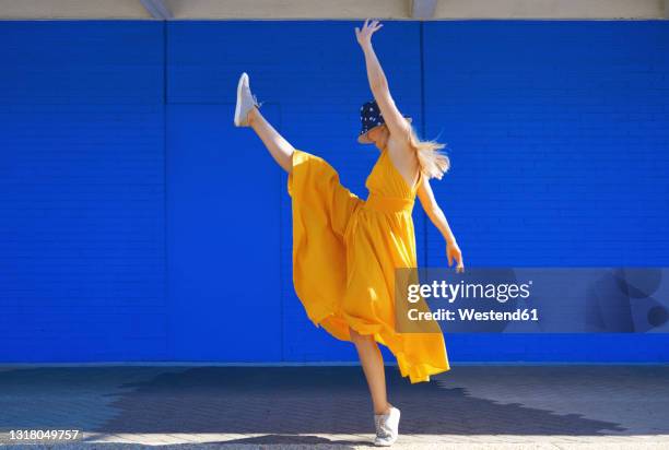 carefree woman dancing by blue wall on sunny day - yellow dress stock-fotos und bilder