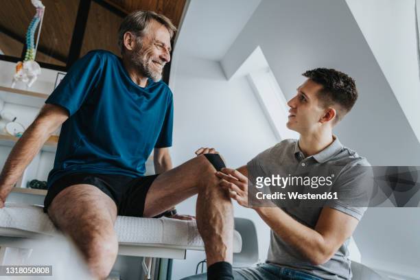 male physical therapist sticking kinesio tape on smiling patient's knee in practice - rehabilitation stock-fotos und bilder