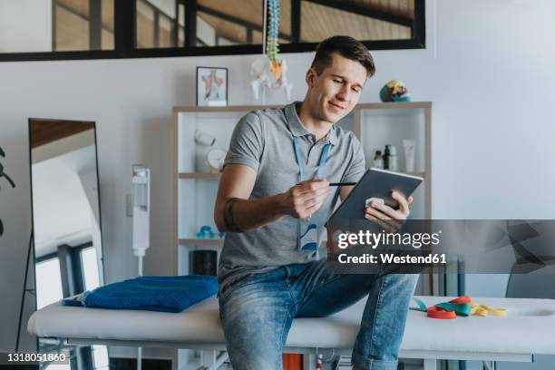 male physiotherapist using digital tablet while sitting on massage table in medical practice - physiotherapist stock-fotos und bilder