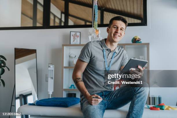 smiling male physiotherapist with digital tablet sitting on bed in medical practice - massage table stock pictures, royalty-free photos & images