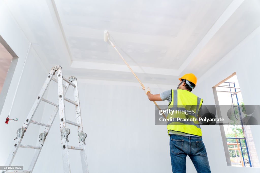 Rear view of asian painter man painting the wall, with paint roller and bucket in an unfinished house, Construction work, isolated on big empty space with ladder