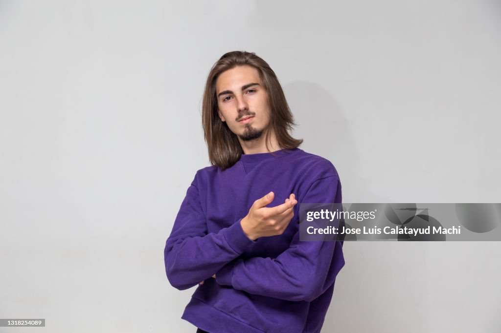 Studio portrait of a young man wearing a purple sweater, with long hair and looking at the camera on a white or gray background