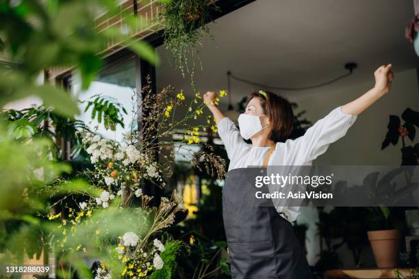 hoopvolle aziatische vrouwelijke bloemist, eigenaar van kleine zakelijke bloemenwinkel met beschermend gezichtsmasker, begint haar werkdag. staand met haar armen uitgestrekt en omhoog kijkend naar de hemel aan de voorkant van de winkel. heropening van bedr - pre opening stockfoto's en -beelden