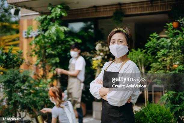 portrait of confident asian woman, owner of small business flower shop, standing at the entrance of shop against colleagues. they are wearing protective face mask to prevent virus. partnership and teamwork. re-opening business after covid-19 concepts - store opening covid stock pictures, royalty-free photos & images