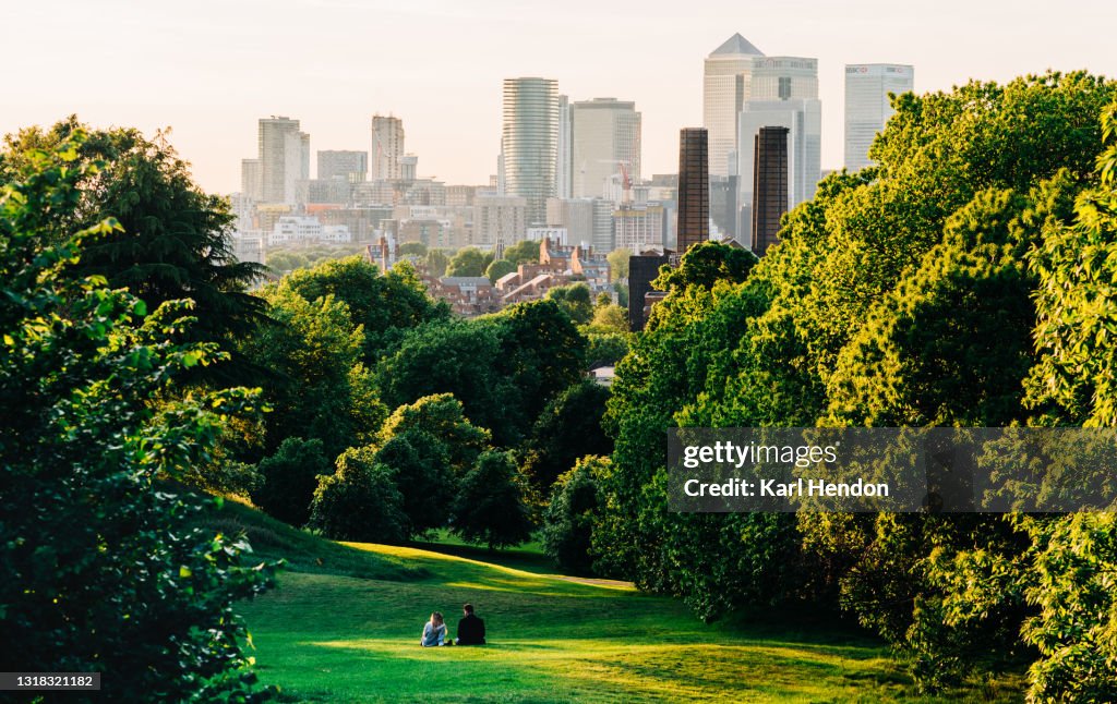 A couple sit in Greenwich Park, London looking the Canary Wharf skyline - stock photo