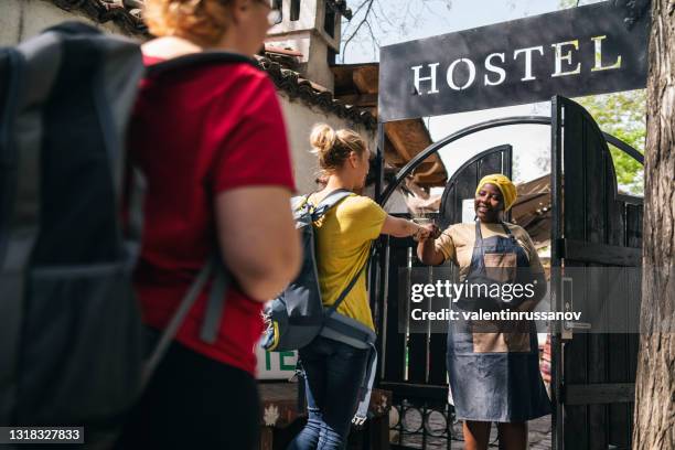 women with backpacks and wearing protective face mask arriving to a city hostel, welcomed by afro female receptionist - hostel stock pictures, royalty-free photos & images