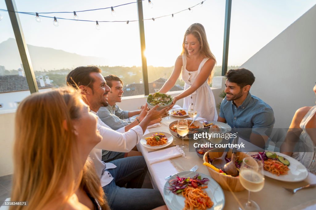 Amigos e familiares passando salada em uma mesa ao pôr do sol.