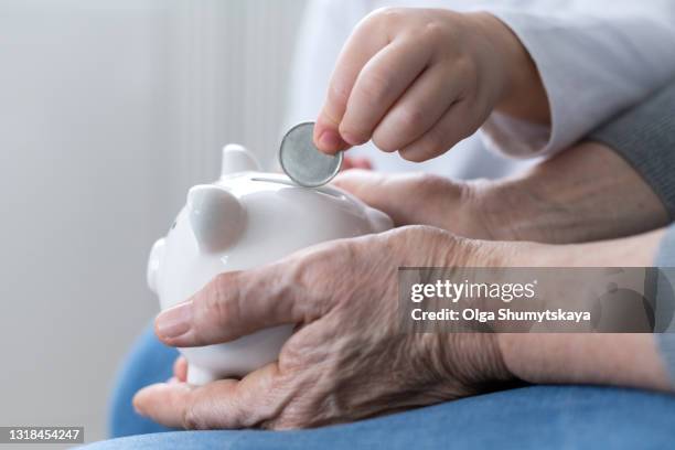 child hand puts a coin in a piggy bank in the hands of an adult elderly - financial support stock pictures, royalty-free photos & images