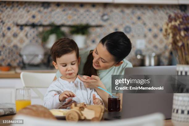 young woman using laptop on the table while her little son having breakfast - balancing act cartoon stock pictures, royalty-free photos & images