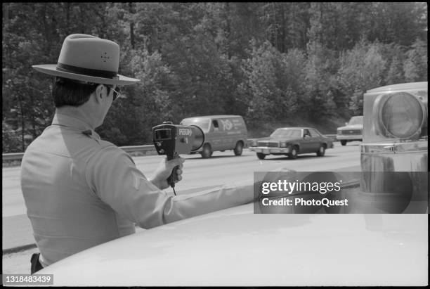 View, from behind, of a Maryland State Police officer as he stands outside his cruisier and uses a radar speed gun to monitor oncoming traffic,...