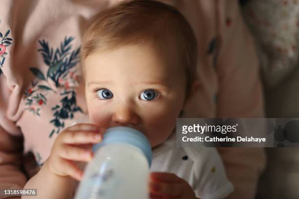 a 1 year old baby girl drinking her baby bottle of milk in the arms of her mum - baby feeding fotografías e imágenes de stock