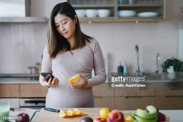 young asian pregnant woman checking nutritional facts and calories intake of assorted fruits with mobile app on smartphone in kitchen at home. healthy diet. eating well. healthy pregnancy eating lifestyle - pregnancy healthy eating stock pictures, royalty-free photos & images
