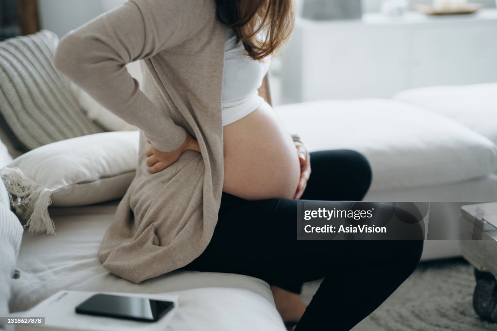 Beskuren skott av asiatisk gravid kvinna som rör vid magen och nedre delen av ryggen, lider av ryggvärk. Graviditetshälsa, välbefinnande koncept