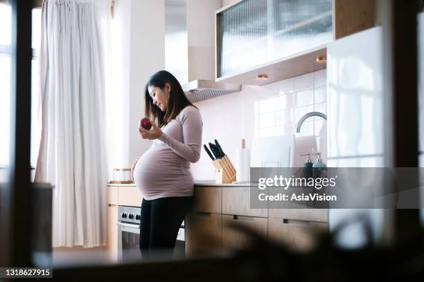 glimlachende jonge aziatische zwangere vrouw wat betreft haar buik, die een verse organische appel in keuken thuis eet. gezonde voeding. goed eten. gezonde zwangerschap die levensstijl eet - asian woman pregnant stockfoto's en -beelden