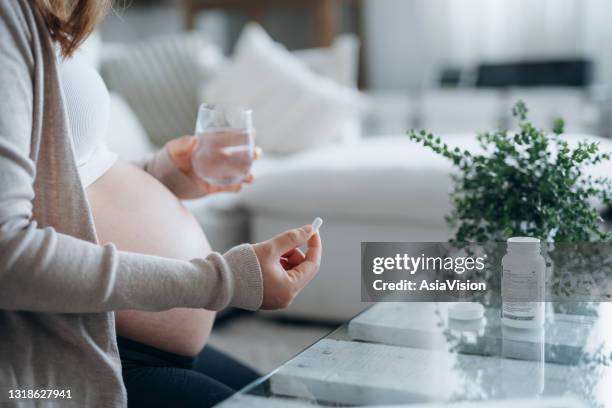 cropped shot of young asian pregnant woman sitting on sofa in living room at home, taking medicine in hand with a glass of water - folic acid stock pictures, royalty-free photos & images