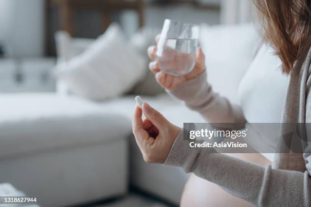 cropped shot of young asian pregnant woman sitting on sofa in living room at home, taking medicine in hand with a glass of water - medicare supplement stock pictures, royalty-free photos & images