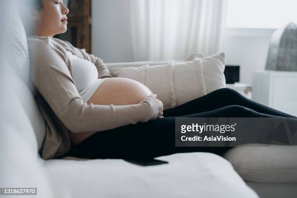 asian pregnant woman with her eyes closed touching her belly, relaxing on sofa at cozy home. expecting a new life, wellbeing, healthy pregnancy lifestyle - prenatal care stock pictures, royalty-free photos & images