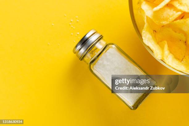 crispy golden fried fat potato chips in a glass bowl or plate, on a bright yellow background or table. next to the salt shaker. the concept of an unhealthy diet and lifestyle, the accumulation of excess weight. copy of the text space. - salt shaker fotografías e imágenes de stock