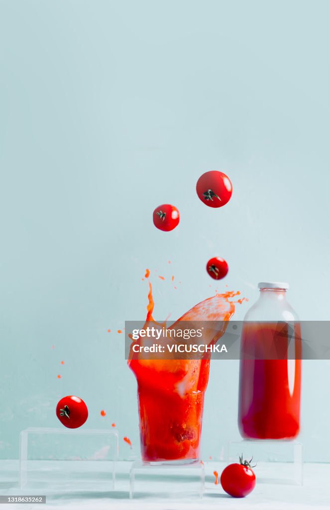Splashing tomato juice in glass, floating tomatoes and glass bottle with juice. Pale blue background. Creative food levitation