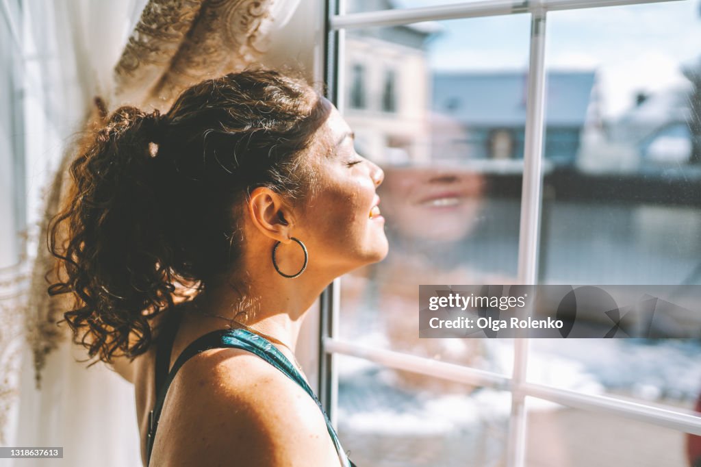 Middle age woman wearing sportswear is standing by the large window and exposing her face to the sun