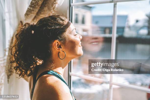middle age woman wearing sportswear is standing by the large window and exposing her face to the sun - luz-solar imagens e fotografias de stock