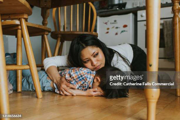 a little boy lies face down on the floor under a table, while a woman comforts him - phobia foto e immagini stock