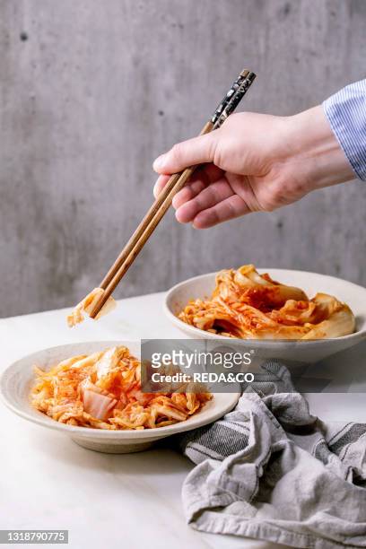 Homemade korean traditional fermented appetizer kimchi cabbage served in ceramic plate over white marble background. Chopsticks in mans hands..