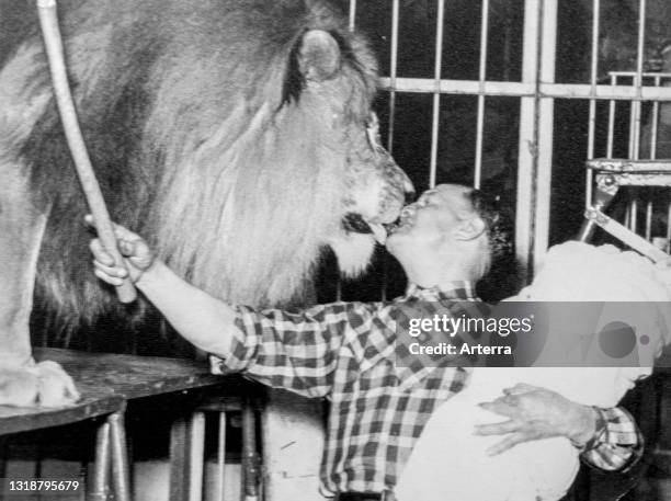 Black and white photograph from the 1950s showing lion tamer giving male lion kiss of death with baby on his arm, tradition among circus performers.