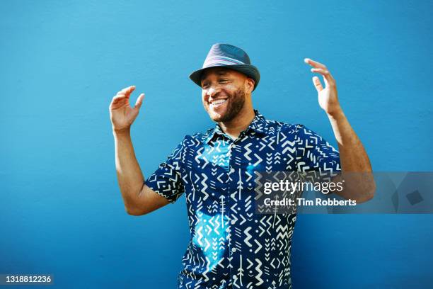 man with arms up, smiling, next to blue wall - cool guy in hat stock pictures, royalty-free photos & images
