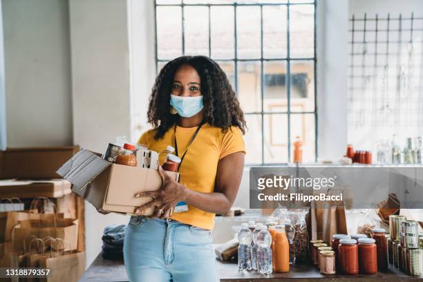 portrait of a mixed race volunteer woman holding a cardboard box of food and drink at the food bank - food staple stock pictures, royalty-free photos & images