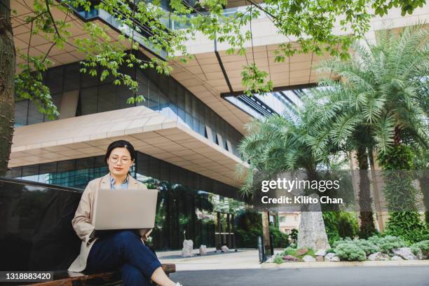 asian woman sitting on a sidewalk using laptop surrounded by luxuriant plants - büropark stock-fotos und bilder