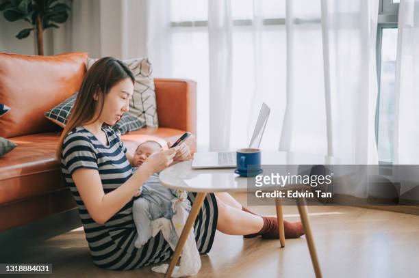 asian chinese beautiful woman working from home using laptop carrying her baby boy in living room - stay at home mum stock pictures, royalty-free photos & images