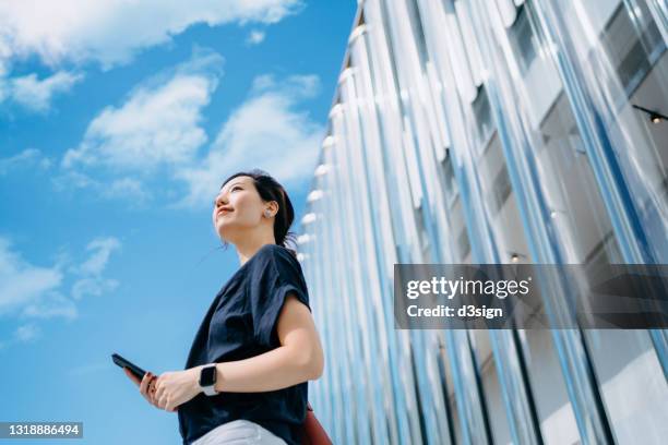 low angle portrait of confident young asian businesswoman with smartphone at downtown district in city, looking up to sky. standing against contemporary corporate buildings. business on the go. girl power. female leadership and determined to success - low confidence stock pictures, royalty-free photos & images
