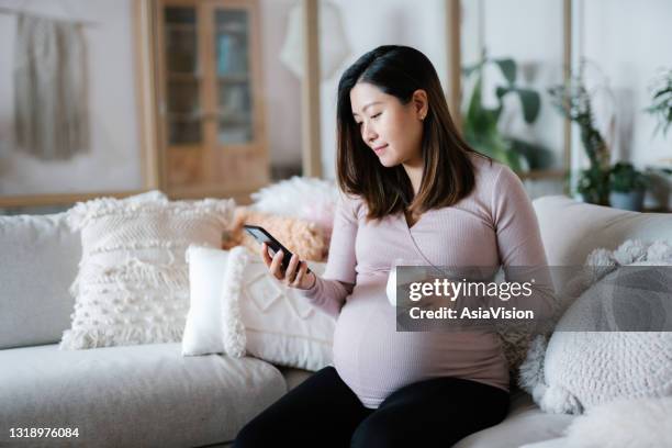 beautiful young asian pregnant woman relaxing on sofa in the living room at cozy home. drinking a glass of fresh milk and using smartphone. wellbeing, healthy eating lifestyle during pregnancy - vitamins and minerals stock pictures, royalty-free photos & images
