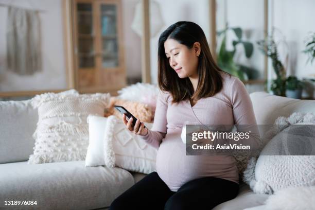 beautiful young asian pregnant woman relaxing on sofa in the living room at cozy home. drinking a glass of fresh milk and using smartphone. wellbeing, healthy eating lifestyle during pregnancy - vitamins and minerals imagens e fotografias de stock