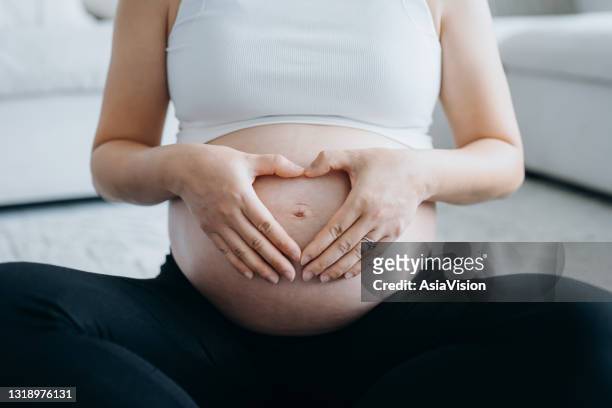 close up mid-section of young asian pregnant woman sitting crossed-legged at home. holding her belly and making a heart shape. expecting a new life, mother's love, staying fit and healthy during pregnancy - human uterus stock pictures, royalty-free photos & images