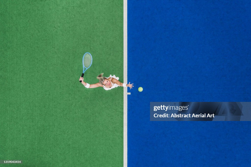 Drone shot looking down on a female about to hit a tennis serve on a two-toned, astroturf, tennis court, England, United Kingdom