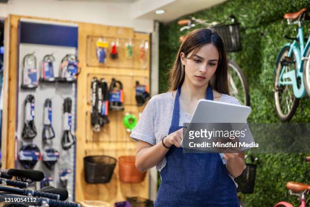 porträt einer jungen ladenbesitzerin, die digitales tablet hält, während sie zwischen fahrrädern steht - sportgeschäft stock-fotos und bilder
