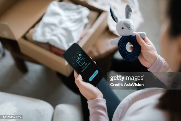 over the shoulder view of asian pregnant woman relaxing on sofa, shopping online with smartphone. she has unboxed a delivery package of baby clothing and toys and holding a bunny toy rattle. time to get some baby essentials for the unborn baby! - baby rattle stock pictures, royalty-free photos & images