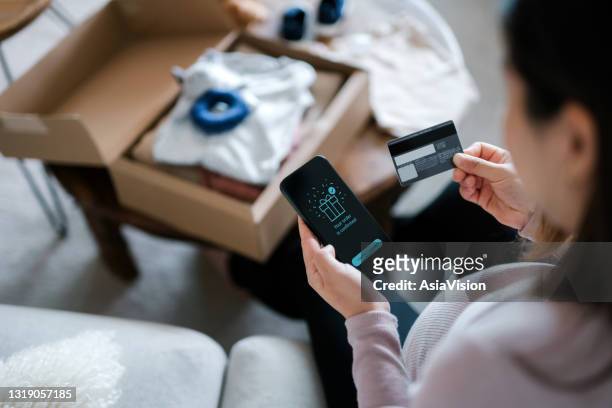 over the shoulder view of asian pregnant woman relaxing on sofa, shopping online with smartphone and making mobile payment with credit card. a delivery package of baby clothing and toys on coffee table. time to get some baby essentials for the unborn baby - shopping online stock pictures, royalty-free photos & images