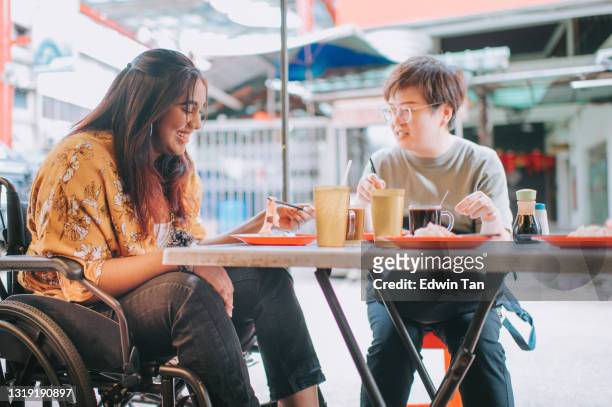 aziatische indische vrouw met handicap op rolstoel die van straatvoedsel in bloemblaadjesstraat met haar chinese vrouwelijke vriend geniet - rolstoelvriendelijk stockfoto's en -beelden